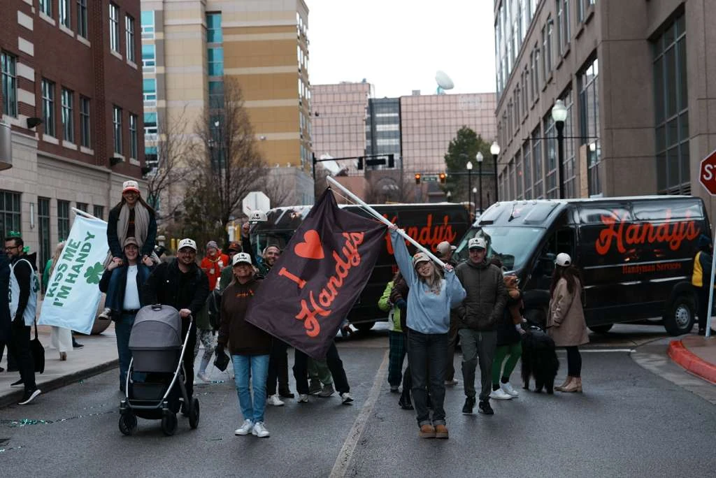 The Handy's crew marching in a parade, waving an I love Handy's flag, with Handy's vans behind